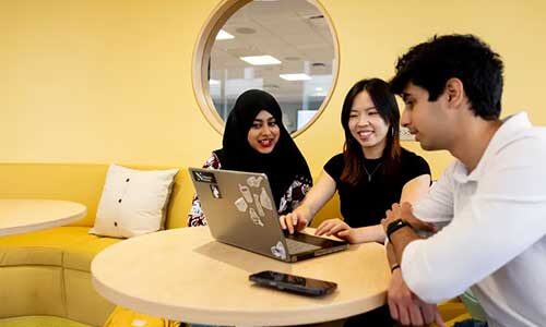 Three students look at a laptop on a small table while sitting in a lounge