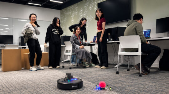 Six students watch a robot they designed move across a floor