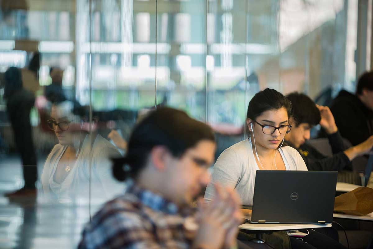 Students sit at individual desks in the West Village H 102 lab. In the center of the image, a student works on a laptop. Her reflection is visible in the glass wall to the left.