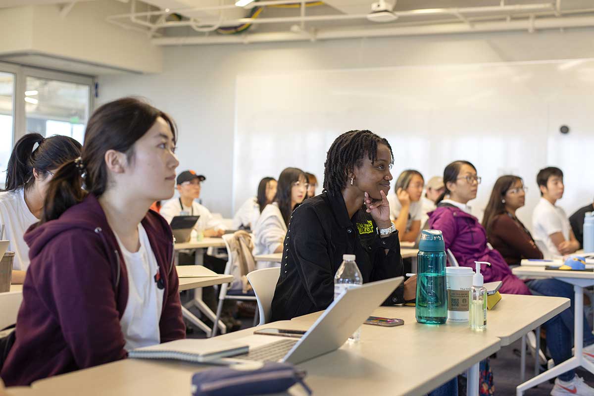 Twelve students sitting in rows of long tables view a class presentation.