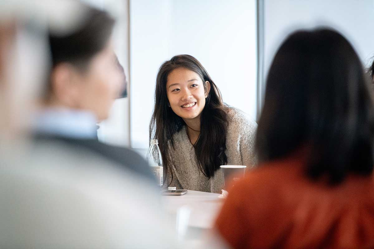 A student sitting at a table in a classroom smiles while listening to a conversation. The backs of two students' heads are visible in the foreground.