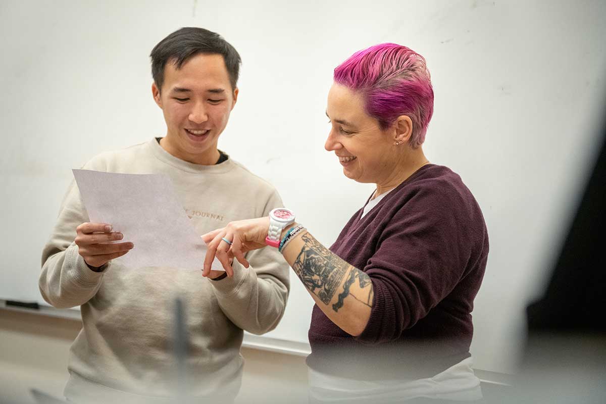 A student (left), holds a piece of paper as faculty member Laney Strange (right) points at it. There is a white board in the background.