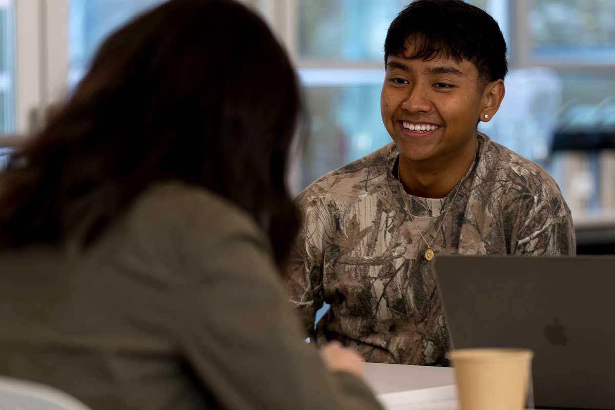 In Seattle, one student sitting at the far side of a table smiles while talking to someone sitting on the other side of the table.