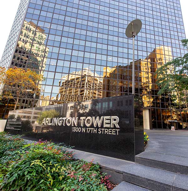 A photo of the Arlington Tower sign with the building rising in the background. The building's glass exterior reflects the surrounding buildings.