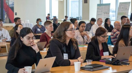 CAGI attendees sit at rows of long tables in a large room