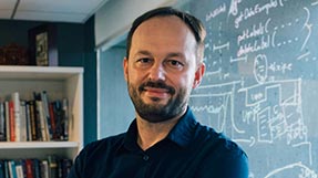 Maciej Kos stands in an office with math equations written on the glass window.