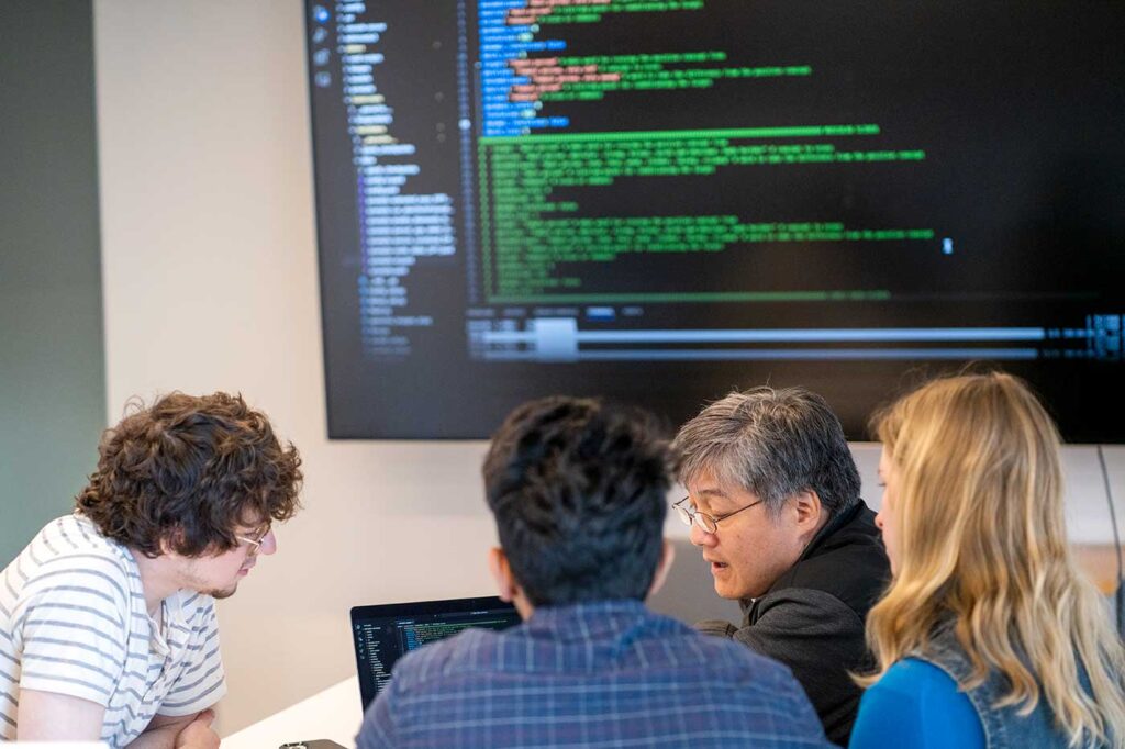 David Bau (second from right), explains code to three students while they sat at a table and view a laptop. The laptop screen's code is projected on the wall in the background