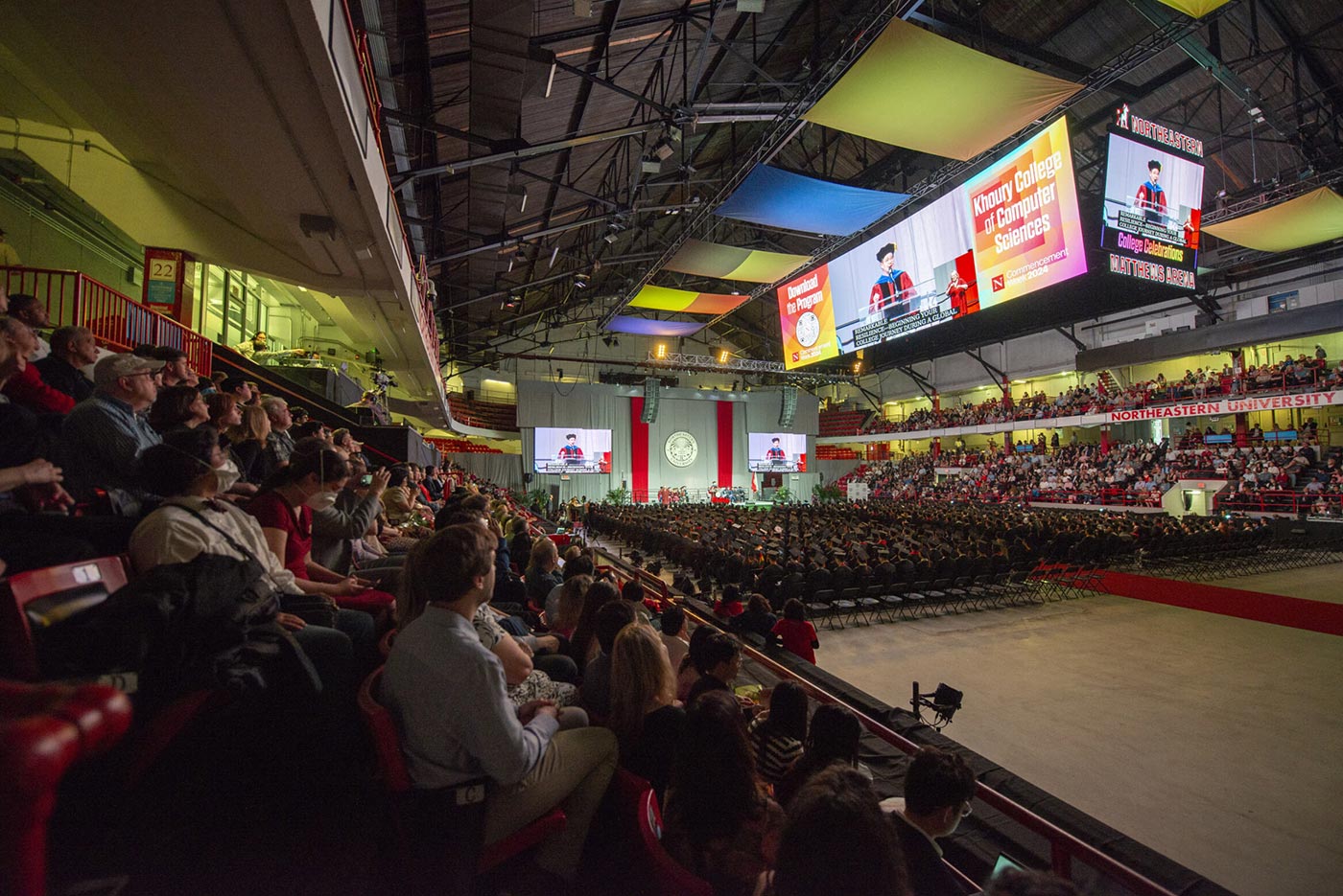 A large crowd in Matthews Arena watches Dean Beth Mynatt's remarks; the video screen is suspended above the arena floor