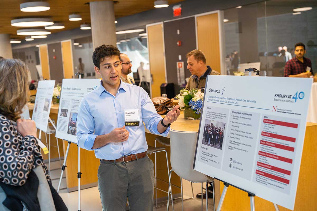 A member of the Sandbox club speaks to a prospective member while gesturing at the club's poster. The poster contains sections that show a photo of the club, the club's mission, partnerships, and impact.