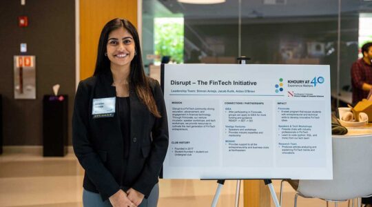 A member of disrupt smiles while standing next to an easel displaying a poster about disrupt. The poster contains sections about the club's mission, history, partnerships, and impact.