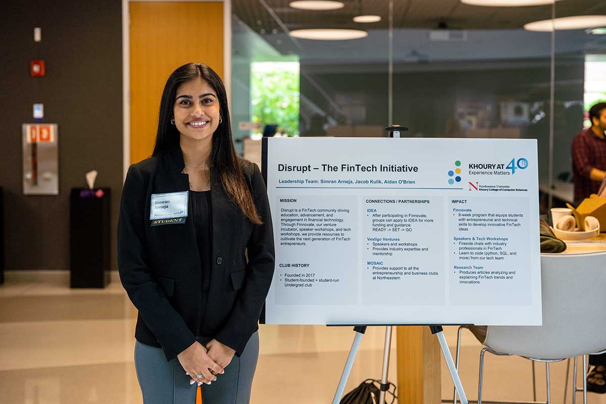 A member of disrupt smiles while standing next to an easel displaying a poster about disrupt. The poster contains sections about the club's mission, history, partnerships, and impact.