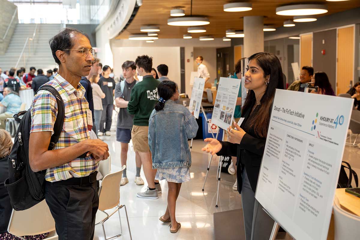 A member of disrupt explains the club's mission to a Khoury College faculty member. The two people are standing next to an easel holding a poster that describes disrupt's mission.