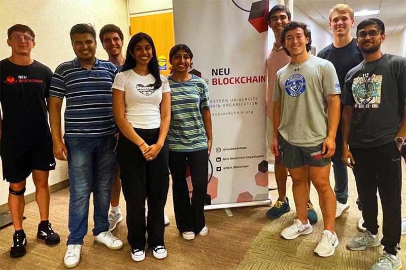 Nine members of NEU blockchain stand around the club's poster in a classroom on Northeastern's campus. Five students are standing to the left of poster and four students are standing on the right.