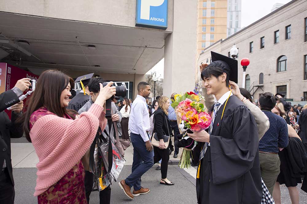 A student wearing a cap and gown poses for a photo while a family member holds a camera