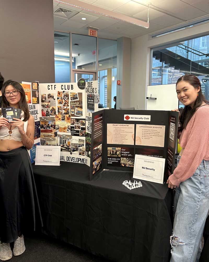 Two members of the CTF club stand in front of their club's table at a club fair. One student is holding up post cards that allow students to register for the club. The other student poses in front of the club's poster.