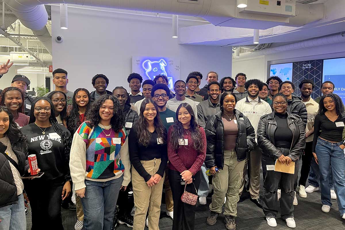 About 35 members of ColorStack pose for a photo in a Northeastern classroom. There is a piece of art with a neon dog head hanging on the wall in the background.