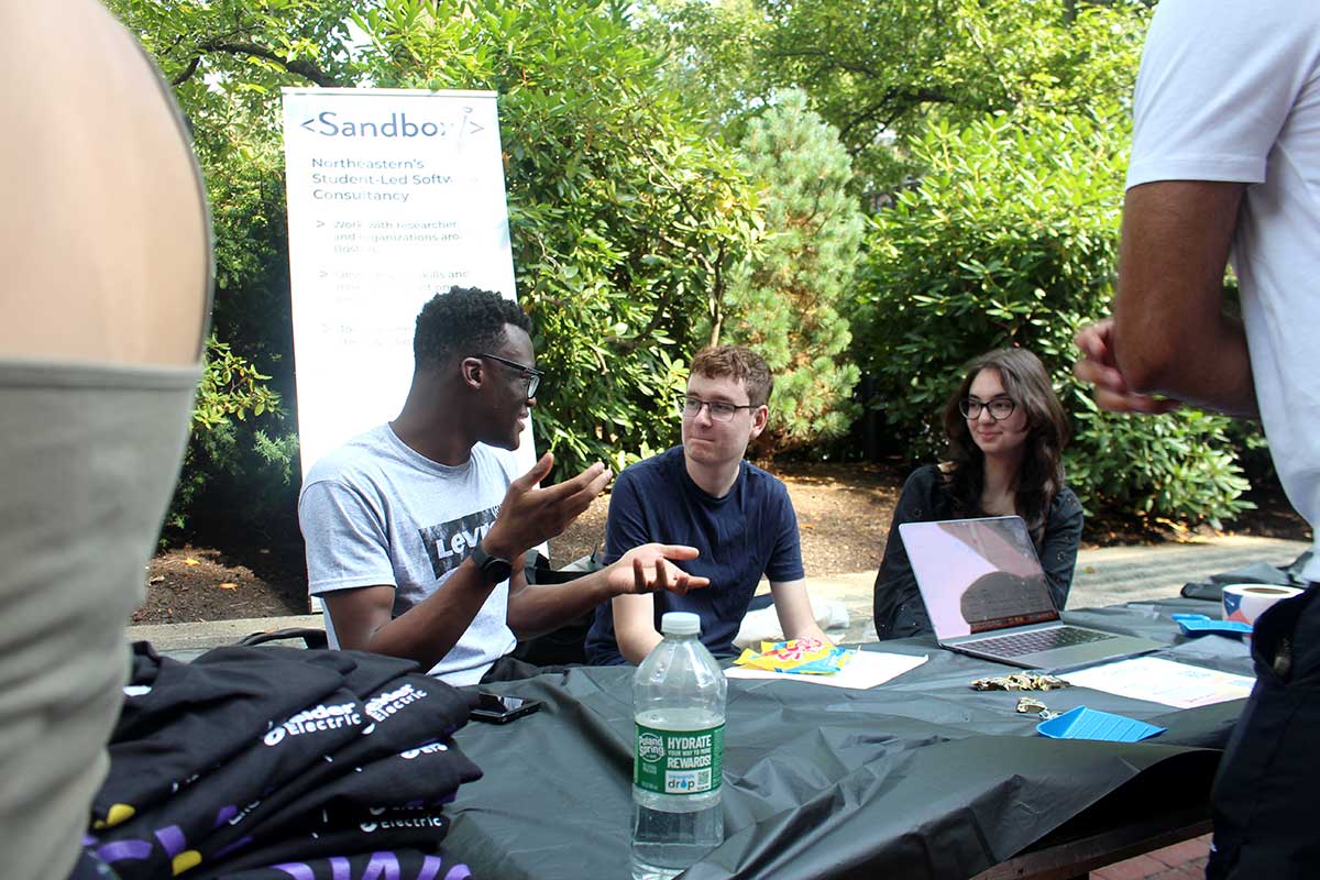 Three members of the Sandbox student club sit behind a table covered by a black tablecloth in a Northeastern courtyard during a club fair. Their club's poster is displayed behind them.