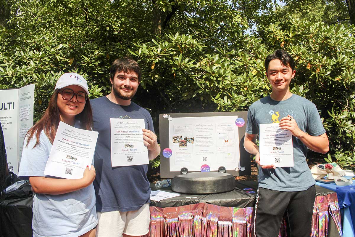 Three student members of code4community pose while holding flyers that describe their club. In the background, there is a table with the club's poster displayed.