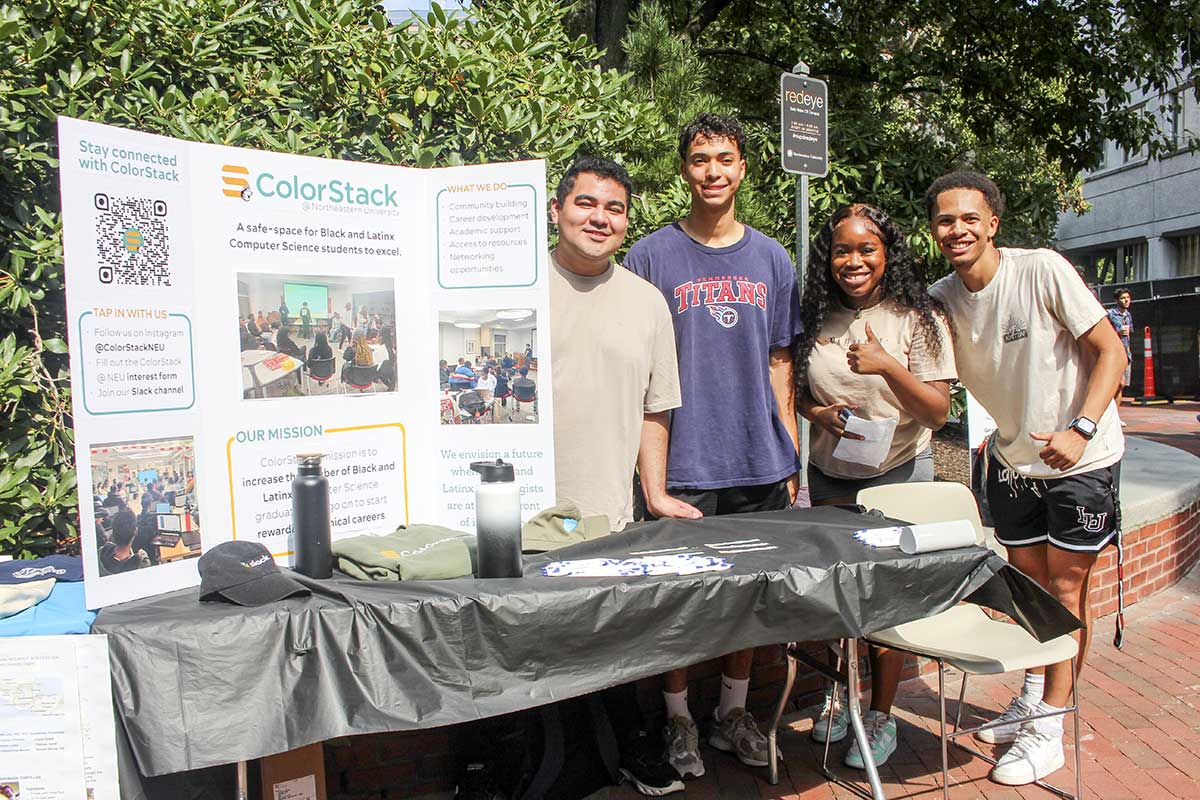 Four members of the ColorStack club stand behind a table while smiling. The table has two water bottles, a hat, a t-shirt, and a poster that describes the club.