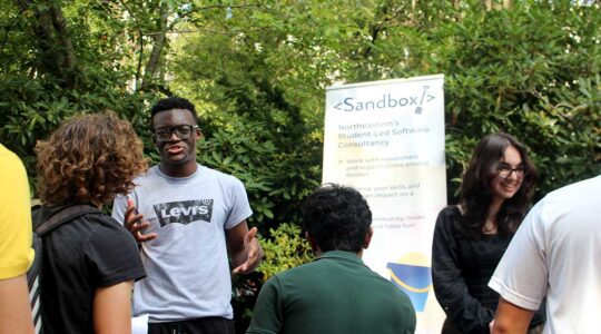 A student member of Sandbox talks about the club to three students.