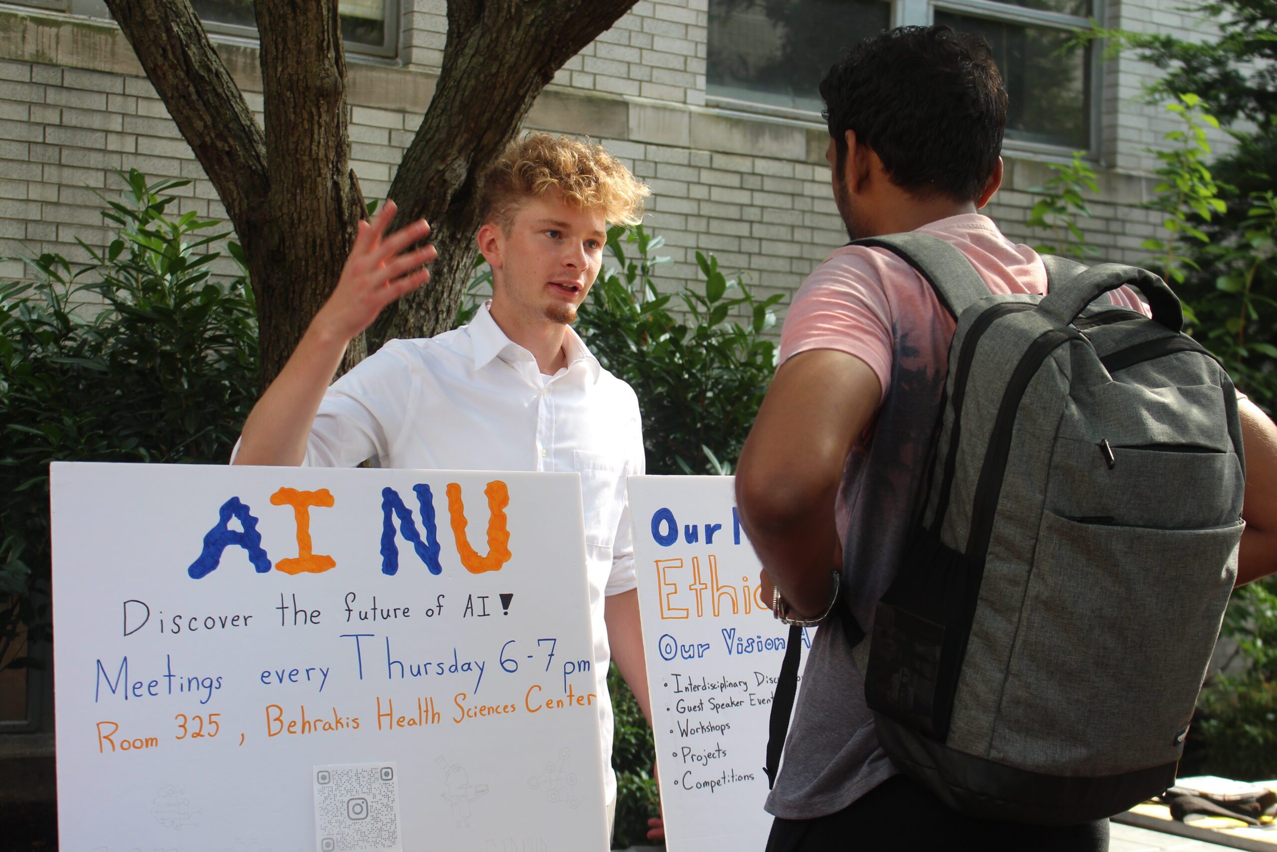 A student standing behind a poster that says "AI at NU" talks about the Artificial Intelligence club with another student