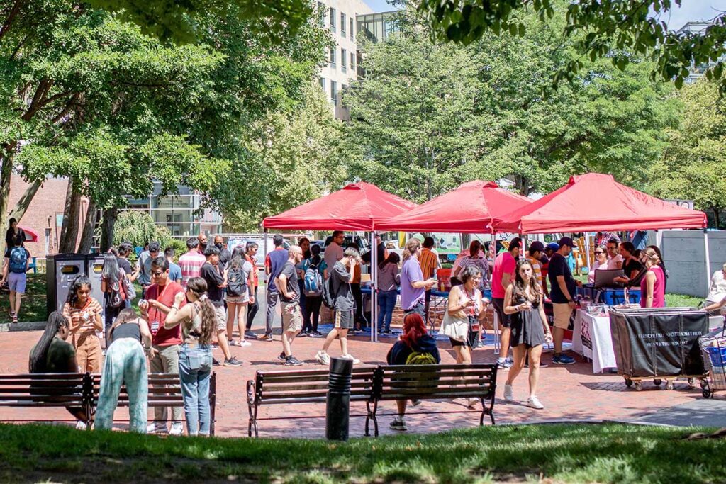 A wide view of the student clubs fair at Northeastern. Three red canopy tents are set up in a campus plaza shaded by trees. Club members stand behind tables under the tents while students interested in joining clubs ask questions.