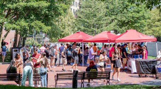 A wide view of the student clubs fair at Northeastern. Three red canopy tents are set up in a campus plaza shaded by trees. Club members stand behind tables under the tents while students interested in joining clubs ask questions.