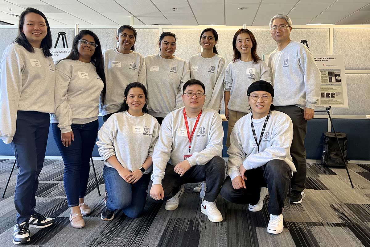 Nine members of the MIG Labs club pose for a photo with the club's advisor, Jeongkyu Lee. Three club members kneel on a rug, and the rest of the people stand behind the first row. Lee is standing in the second row at the far right.