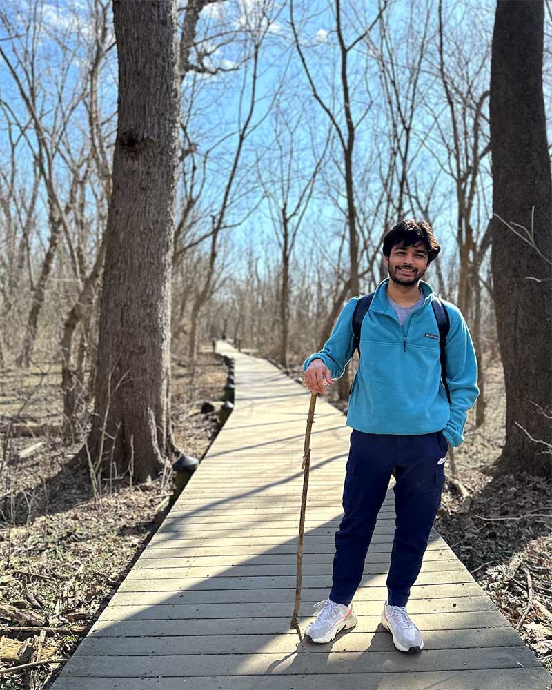 A Khoury student poses on a walking trail in a wooded area near the Northeastern Arlington campus. The student is holding a walking stick and the trail is marked by wooden boards.