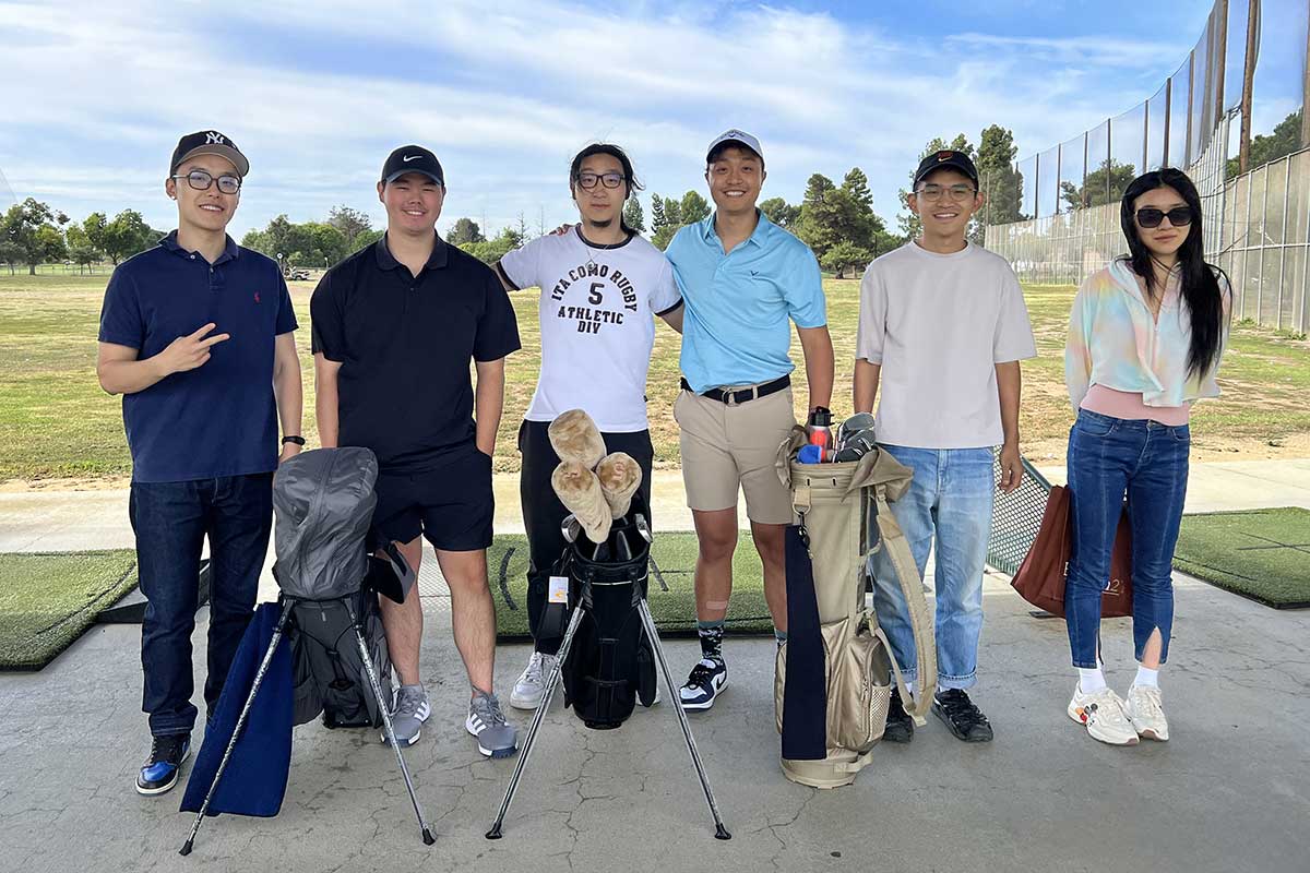 Six members of the Bay Area Golf Club pose for a photo on a sunny day at a local driving range. There are three golf bags standing in front of the club members.