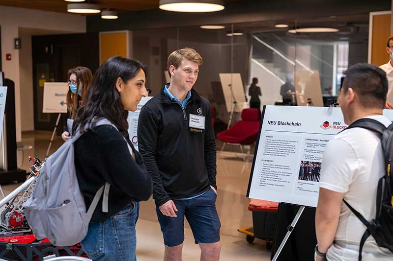 A member of NEU Blockchain stands next to his club's poster, which is displayed on an easel, while two other students view the poster and ask questions about the club.
