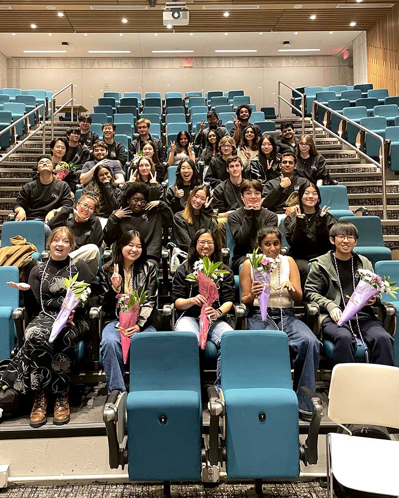 A photo showing about 33 members of Code4Community sitting in a Northeastern lecture hall. The five students sitting in the first row are holding bouquets of flowers.