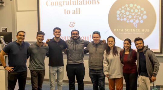 Eight members of the Data Science Hub pose for a photo in front of a projection screen in a classroom.