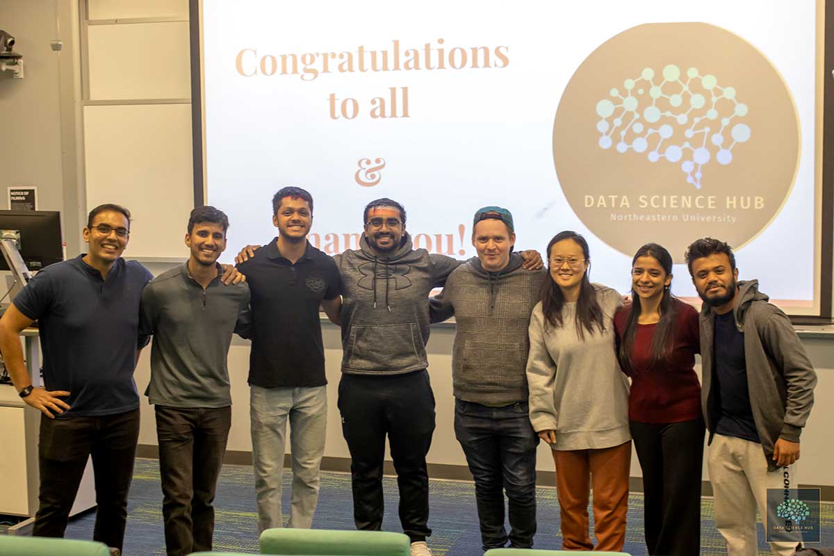 Eight members of the Data Science Hub pose for a photo in front of a projection screen in a classroom.