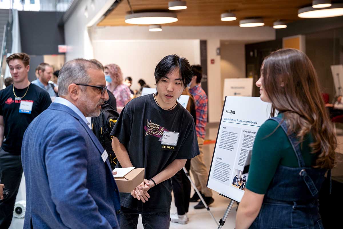 Two members of the firstbyte student club listen to a question being asked by a Northeastern faculty member. The club's poster is visible between the three people.