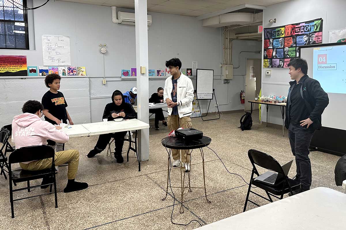 Two members of firstbyte lead a disucssion in a classroom with four middle school students interested in STEM. Three students are seated at tables and another student is standing.