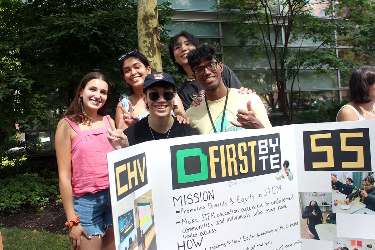 Five members of firstbyte pose for a photo while standing behind their club's poster at a Northeastern club far. The student are smiling and showing peace signs.