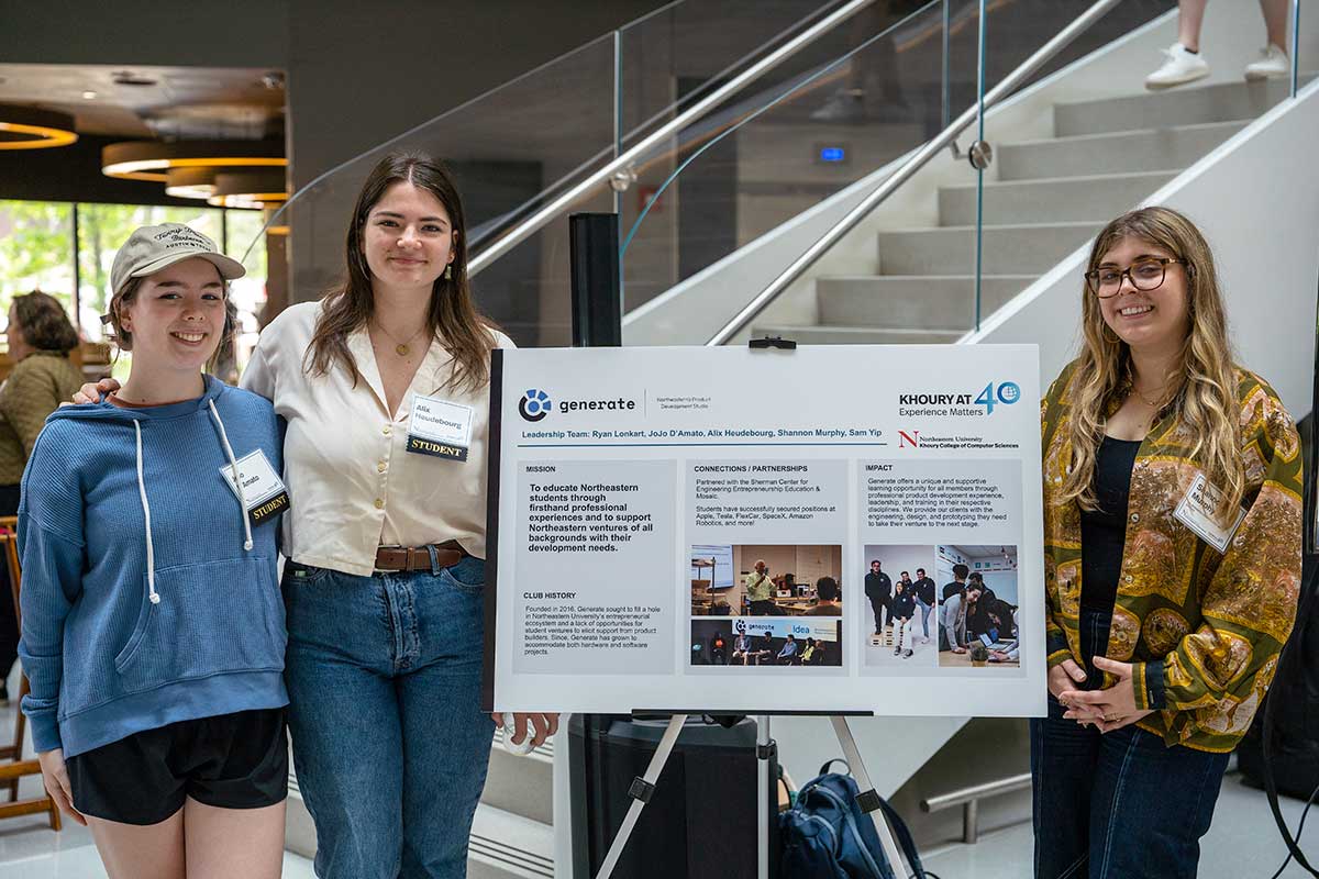 Three members of Generate stand near their club's poster at a club fair in Northeastern's ISEC building. Two students are standing to the left of the poster and one student is standing to the right.