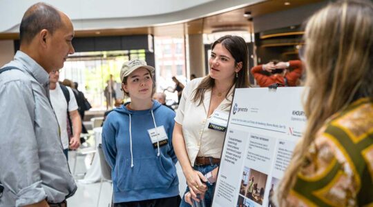 A member of Khoury's Generate student club describes the club to a faculty member. In the background, another club member listens in to the conversation.