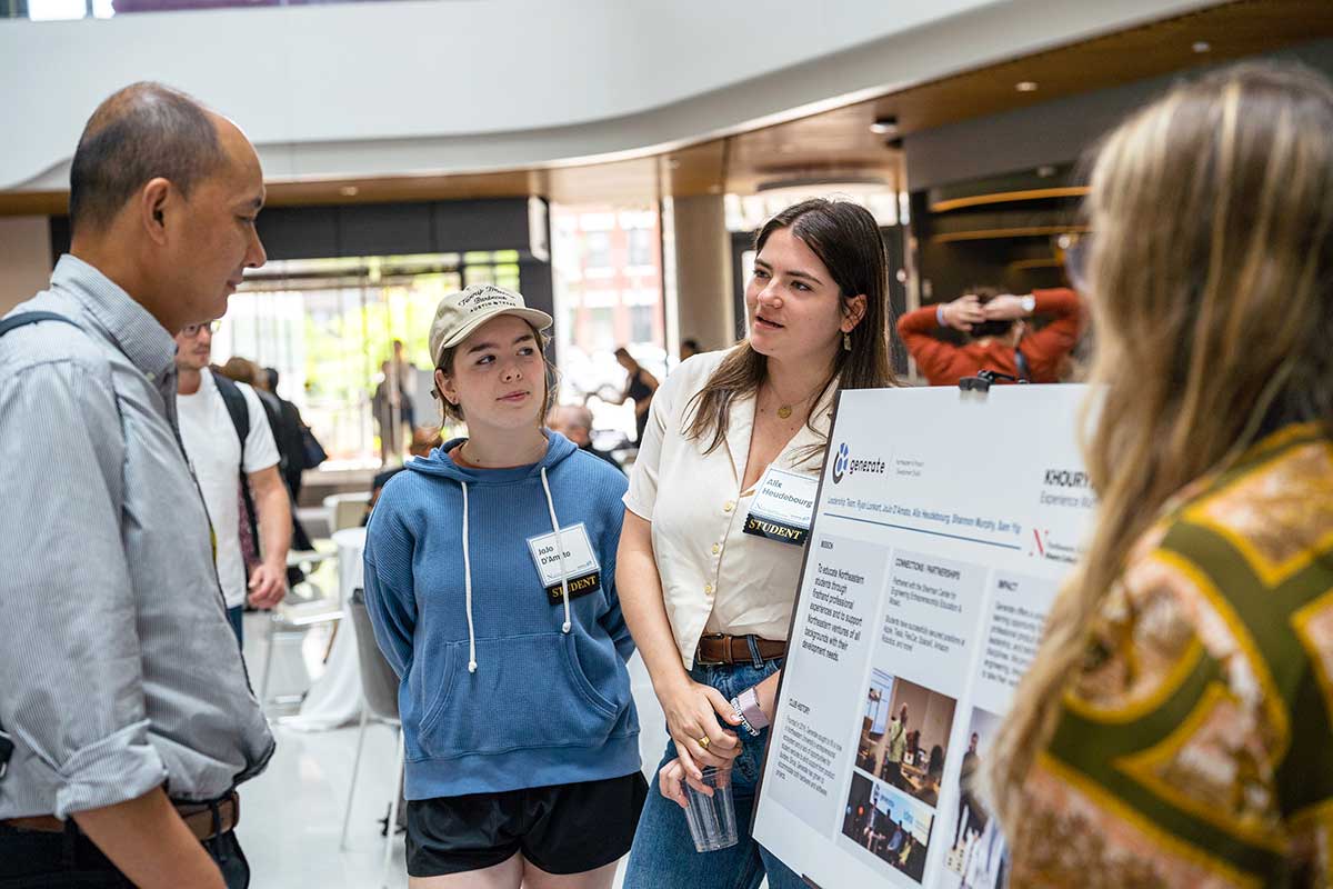 A member of Khoury's Generate student club describes the club to a faculty member. In the background, another club member listens in to the conversation.