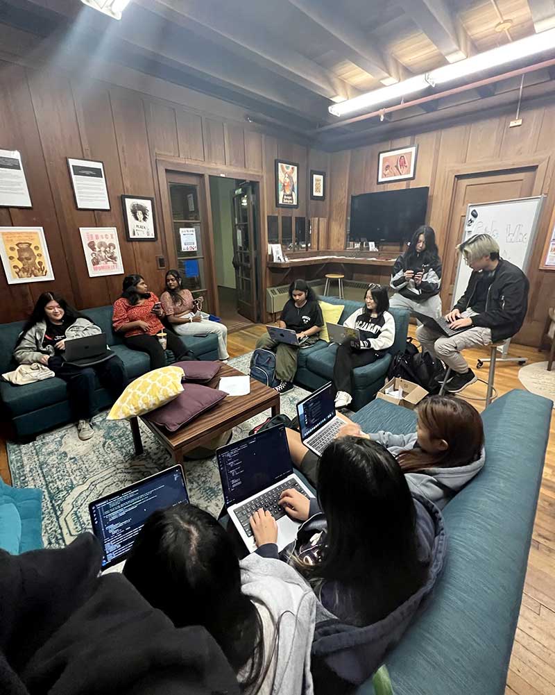 Ten members of Girls Who Code - Oakland sit on couches and chairs while working on their laptops in a room at Northeastern University - Oakland.