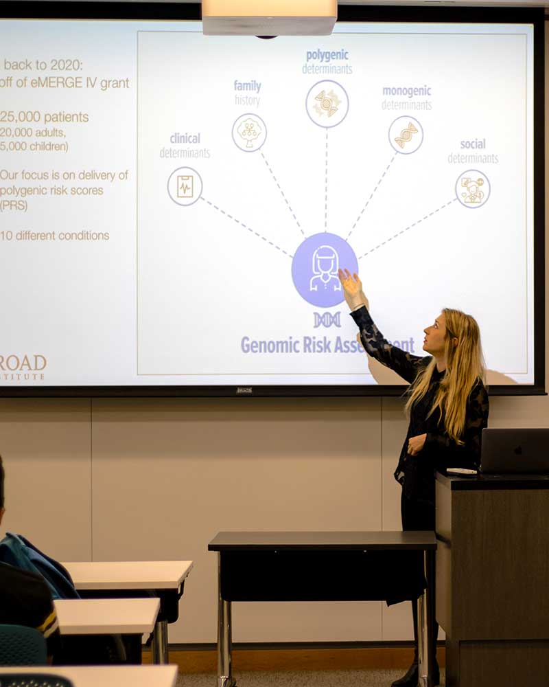 A member of the Google Developer Student Club gives a presentation in a classroom. The student is pointing at a projection screen. The screen shows a diagram titled Genomic Risk Assessment.