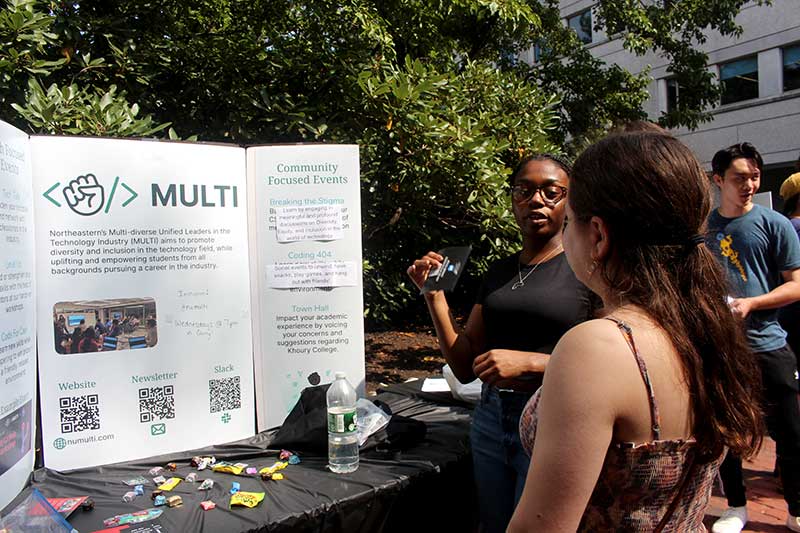 Two students stand in front of a table discussing MULTI's club activities while looking at the club's poster.