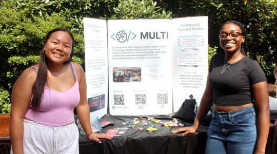 Two members of MULTI pose for a photo at a Northeastern club fair. The students are standing in front of a table that displays the club's poster.