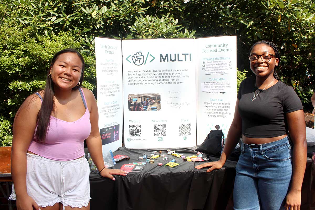 Two members of MULTI pose for a photo at a Northeastern club fair. The students are standing in front of a table that displays the club's poster.