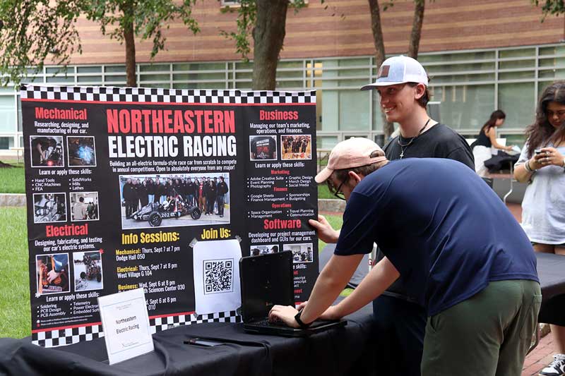 The Northeastern Electric Racing club's poster is displayed on a table. One student types at a laptop in front of the poster while another student looks on.