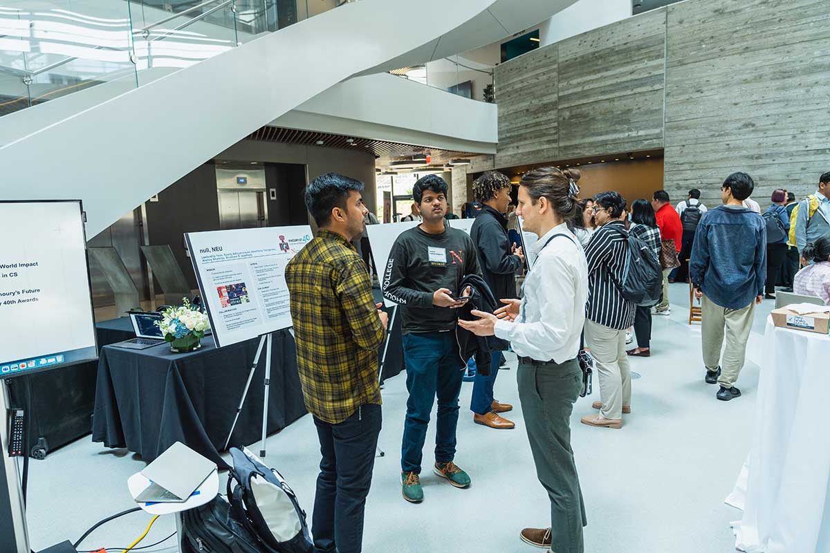 Two members of the null NEU club discuss the club with a Khoury faculty member in the ISEC building's atrium. The club's poster is displayed on an easel behind the students.