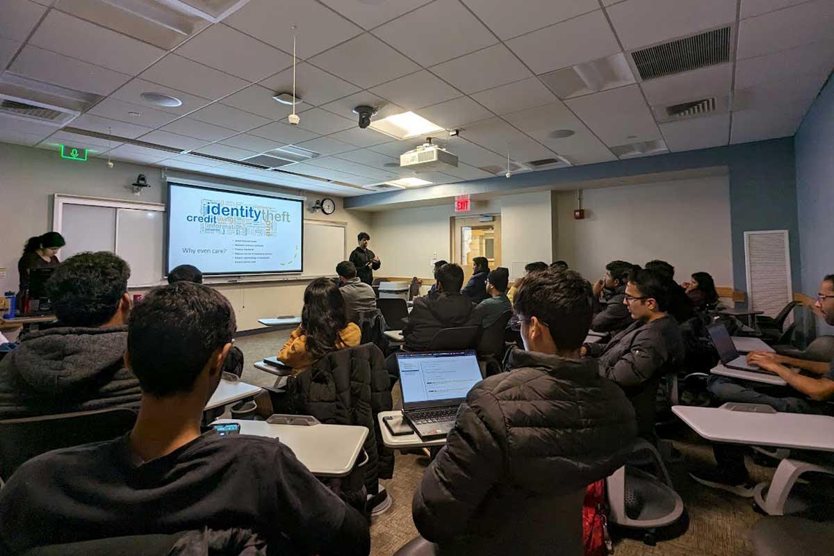 A photo taken from the back corner of a classroom during a null NEU meeting. There are about 20 members sitting at desks viewing a presentation being given by one member standing at the front of the room. The words "identity theft" are visible on the projection screen at the front of the room.