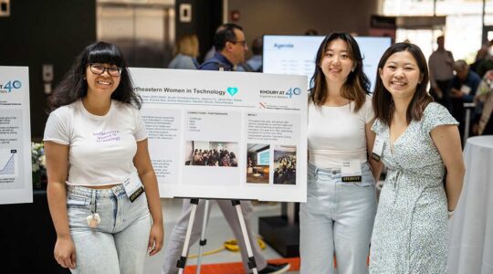 Three students from NU Women in Technology stand adjacent to their club's poster during a club fair inside a Northeastern building. One student stands to the left of the poster and the two other students stand to the right.