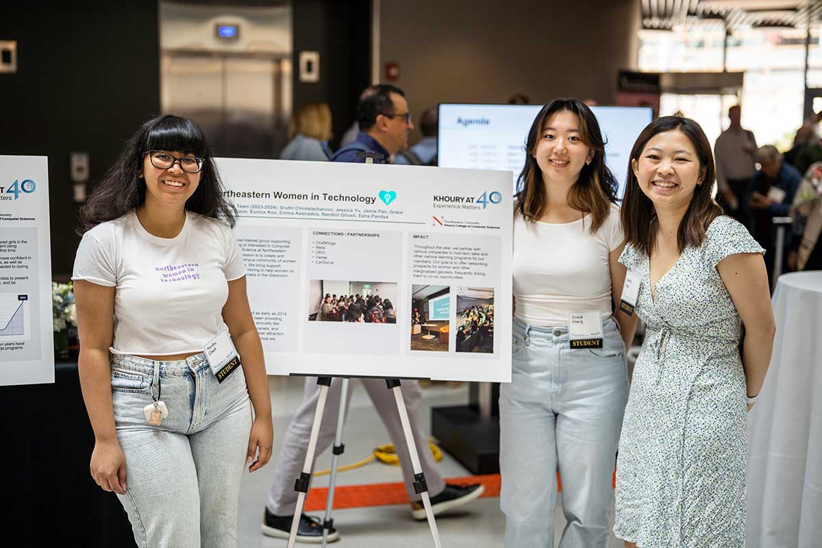 Three students from NU Women in Technology stand adjacent to their club's poster during a club fair inside a Northeastern building. One student stands to the left of the poster and the two other students stand to the right.